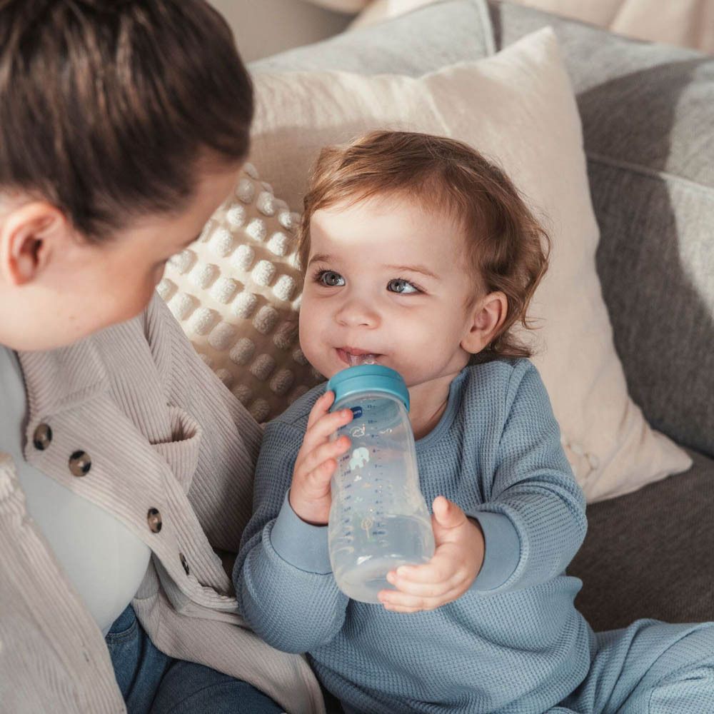 Ein Baby trinkt aus einer transparenten Trinkflasche mit blauem Deckel. Eine Frau hält die Flasche.