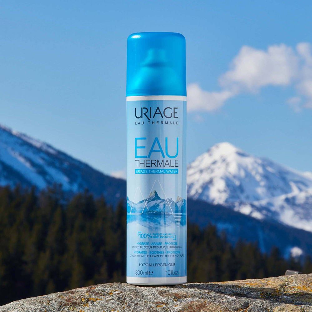 Spray Uriage Eau Thermale davanti a un paesaggio montano. Flacone blu con etichetta bianca.