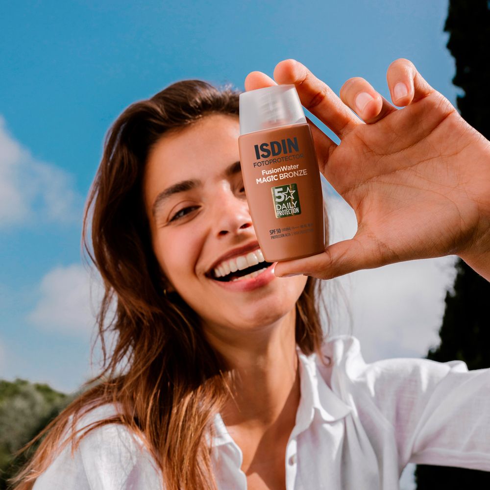 Frau hält eine Sonnencreme-Flasche. ISDIN, Fusion Water, Magic Bronze, SPF 50. Lächelt. Blauer Himmel.