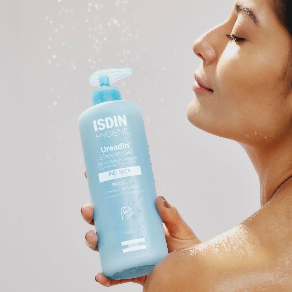 Donna tiene flacone azzurro con dosatore. Scritta: ISDIN Hygiene Ureadin Shower Gel. Spruzzi d'acqua.