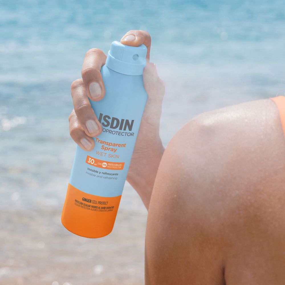 Person hält eine blaue Sprühflasche. ISDIN Fotoprotector Transparent Spray Wet Skin SPF30. Sprüht auf die Haut.