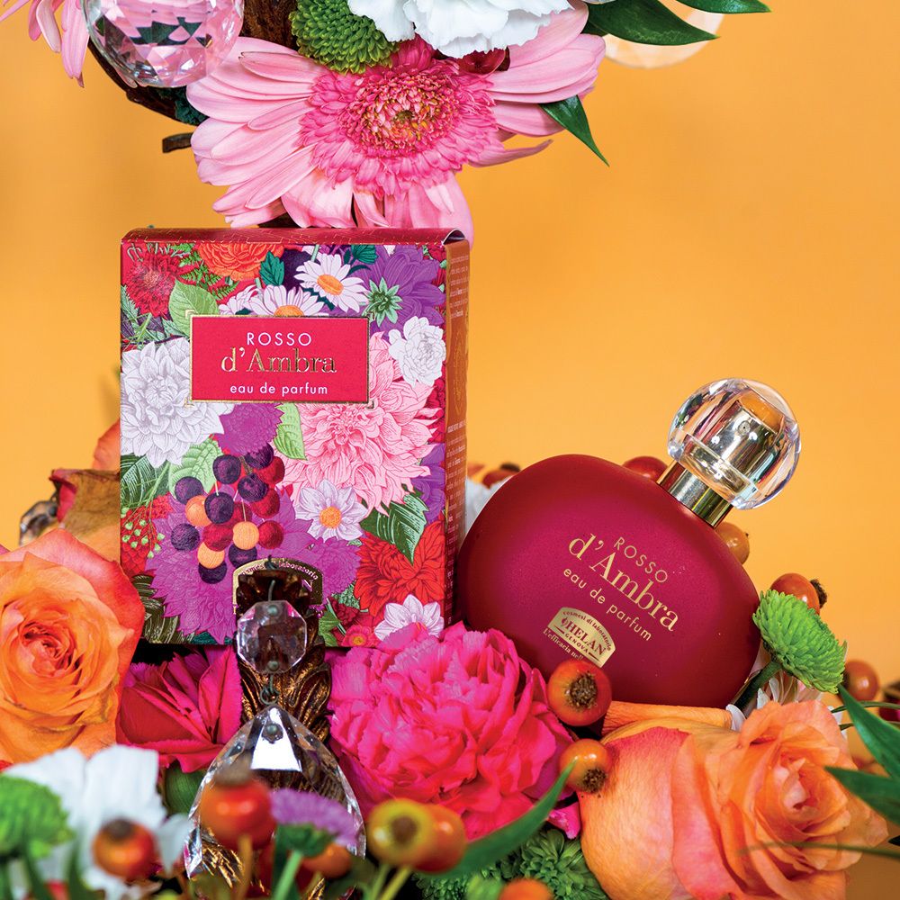 Flakon und Verpackung mit Blumen. Aufschrift: ROSSO d'Ambra, Eau de Parfum. Zusätzliche Flakons. Gelber Hintergrund.
