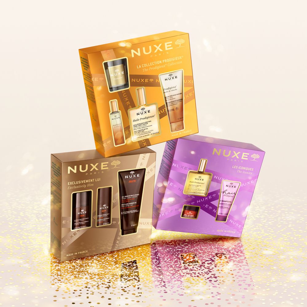 Tre confezioni regalo: oro, marrone e viola. Ogni set contiene diversi prodotti. Logo Nuxe e nomi dei prodotti visibili.
