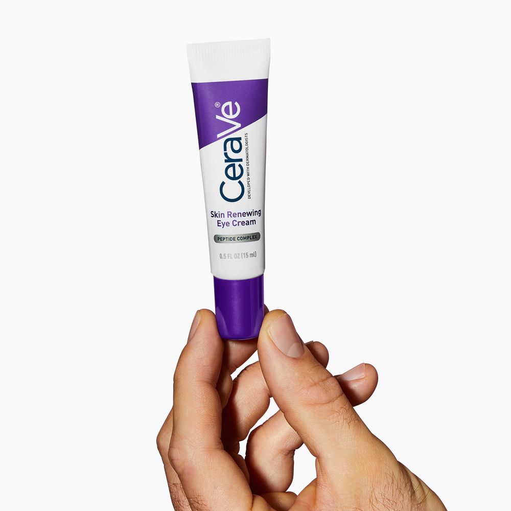 Hand hält CeraVe Skin Renewing Augencreme-Tube. Weiß, lila. Produkt sichtbar. Helle Umgebung.