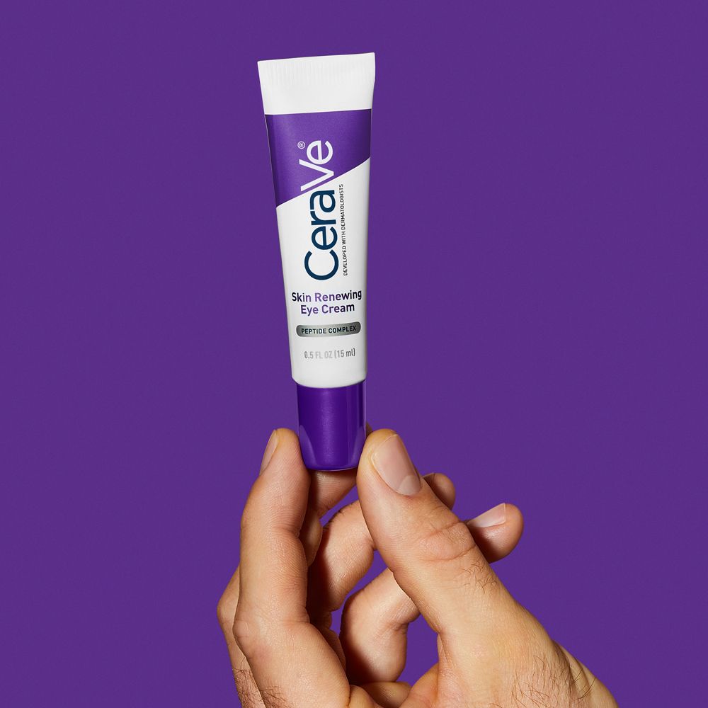 Hand hält CeraVe Skin Renewing Augencreme-Tube. Weiß, lila. Produkt sichtbar. Lila Hintergrund.
