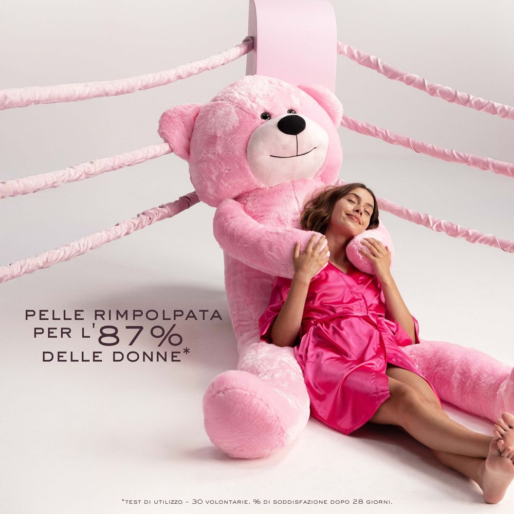 Donna che abbraccia un orso di peluche rosa gigante. Testo: Pelle rimpolpata per l'87% delle donne.
