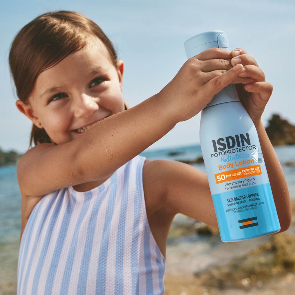 Kind hält eine hellblaue Sprühflasche mit ISDIN-Logo. Strandhintergrund. Kind lächelt.