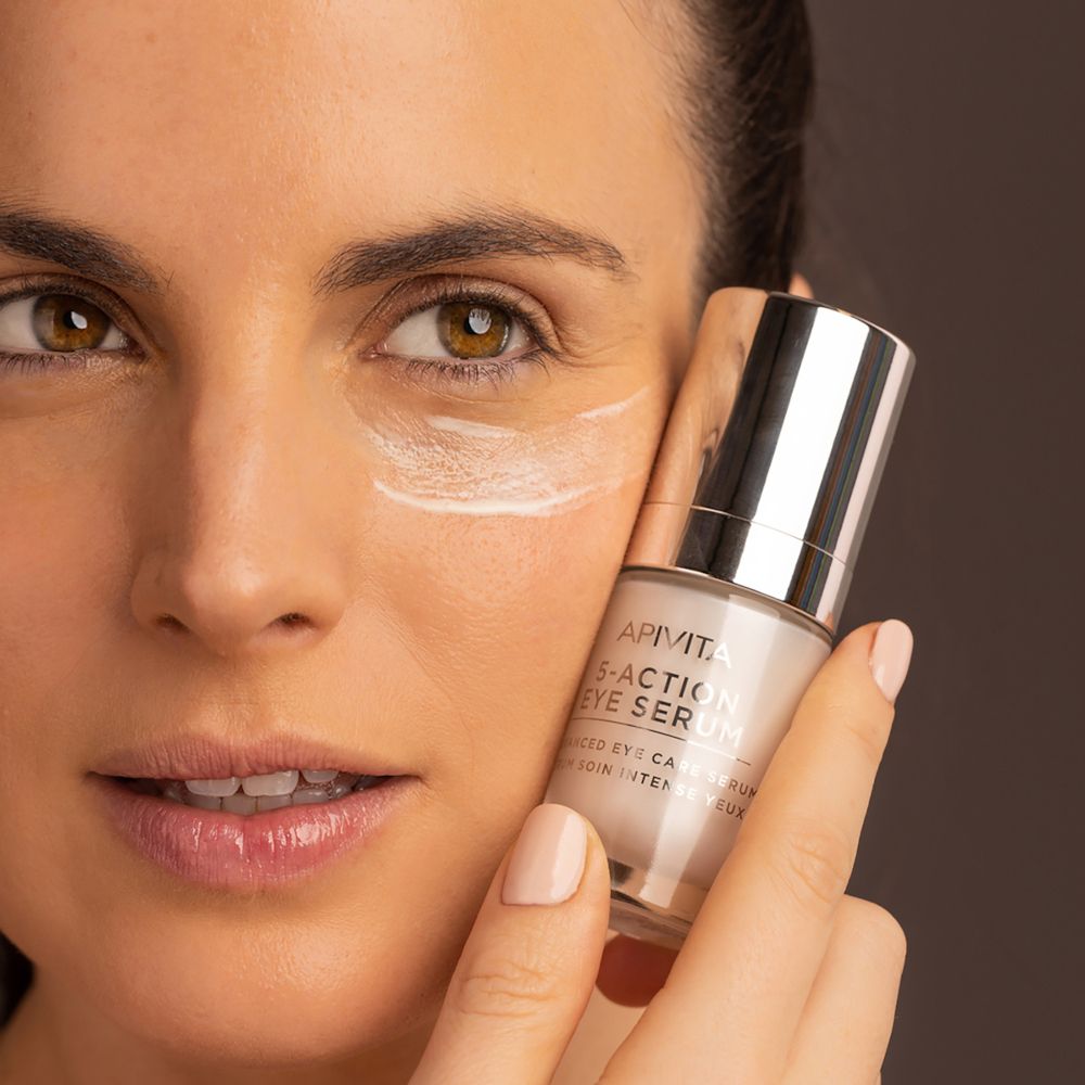 Donna che tiene in mano un flacone di siero. Scritta: APIVITA 5-Action Eye Serum. Crema sulla pelle.