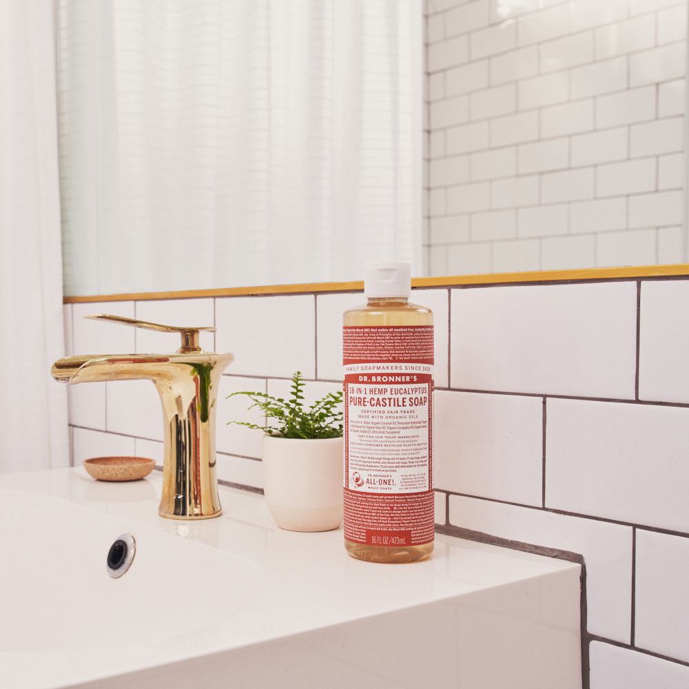 Sapone Dr. Bronner's Eucalyptus vicino al lavandino. Rubinetto dorato, pianta in vaso. Bottiglia con tappo bianco.
