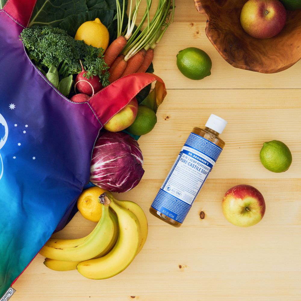 Una bottiglia di sapone liquido è appoggiata accanto a frutta e verdura in una borsa della spesa su un tavolo di legno.