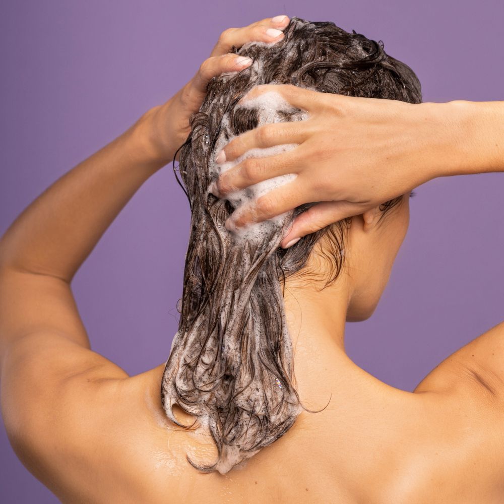 Donna si lava i capelli con shampoo. Schiuma sui capelli. Sfondo: viola.