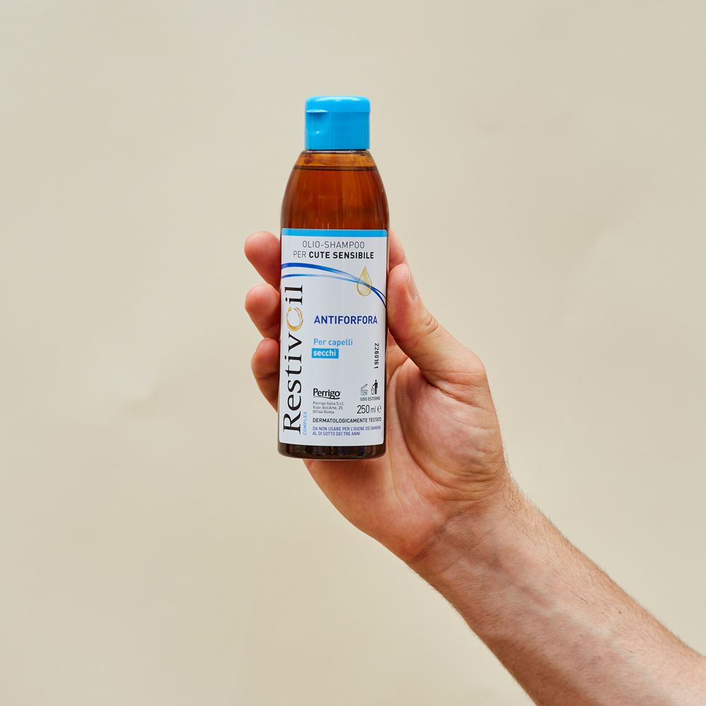 Flacone di shampoo all'olio Restivoil. Bottiglia marrone, tappo blu. Tenuto in mano. 250ml.