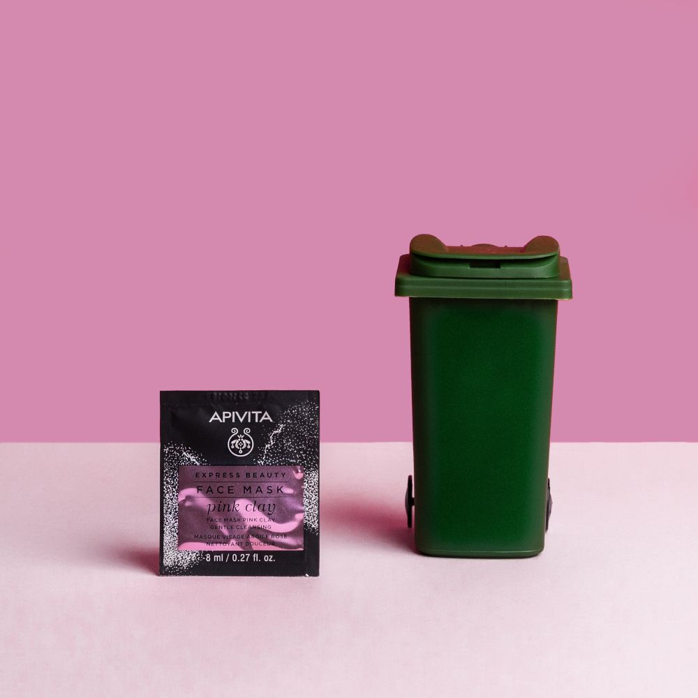 Rosa Hintergrund. Ein Beutel mit APIVITA EXPRESS BEAUTY FACE MASK pink clay neben einem grünen Mülleimer.