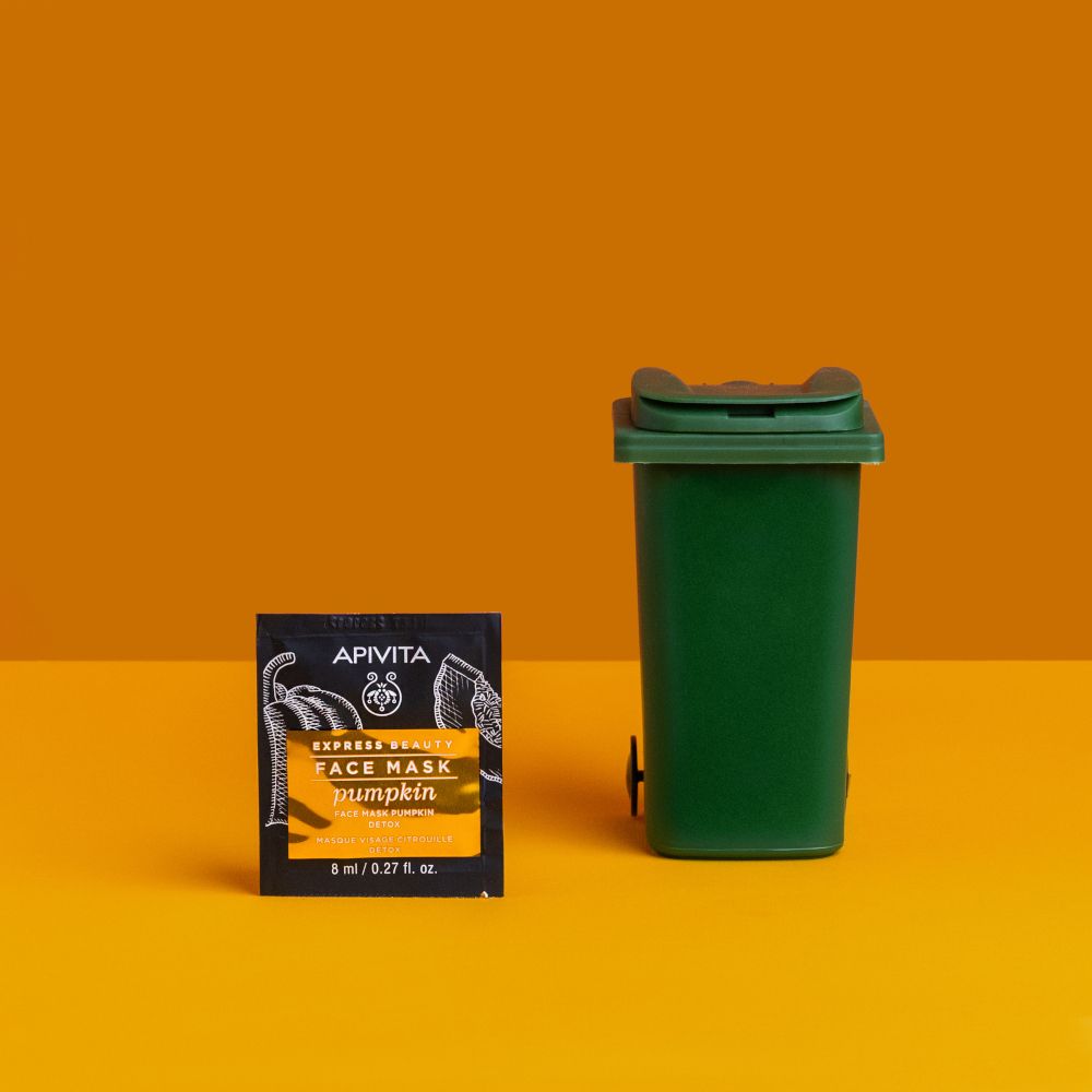 Bustina APIVITA EXPRESS BEAUTY FACE MASK Pumpkin accanto a un bidone verde su sfondo giallo.
