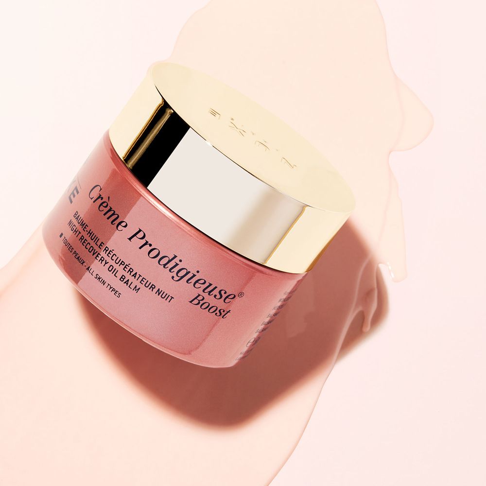 Vaso di crema rosa inclinato con coperchio dorato. Scritta: Crème Prodigieuse Boost, Baume-huile récupérateur nuit.