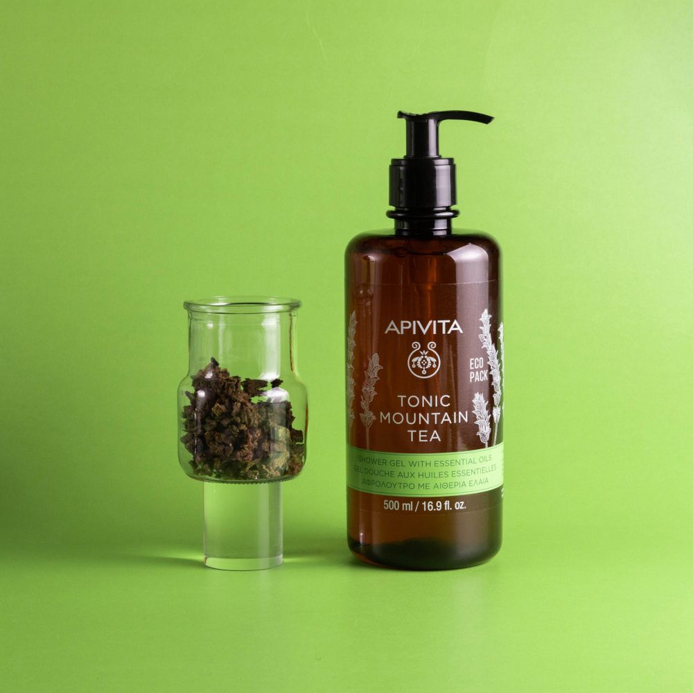 Braune Pumpspendeflasche mit APIVITA TONIC MOUNTAIN TEA Aufschrift und ein Glasbehälter mit Bergtee-Blättern.