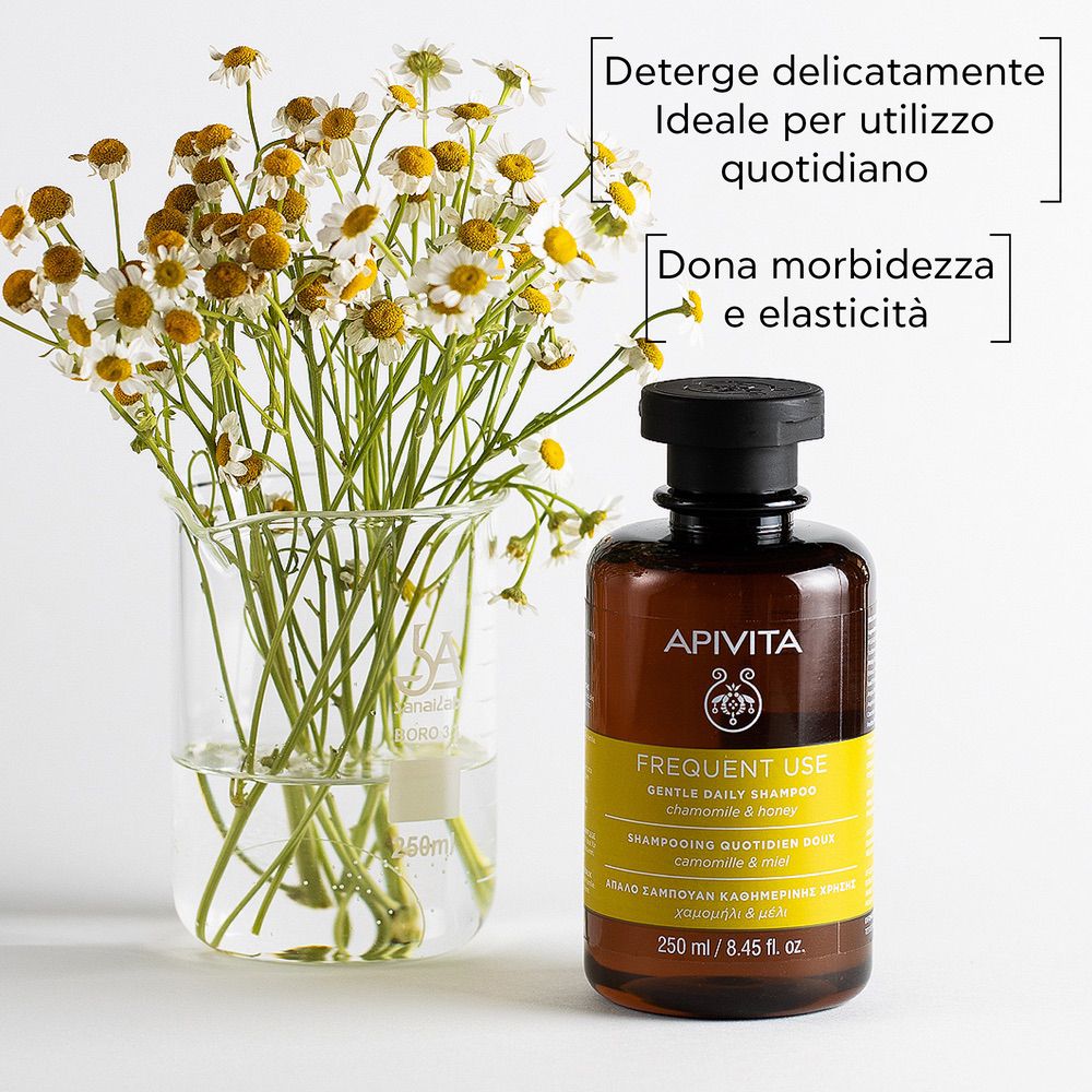 Flacone di shampoo accanto a fiori di camomilla in un bicchiere. Marchio: APIVITA. Testo sull'etichetta.