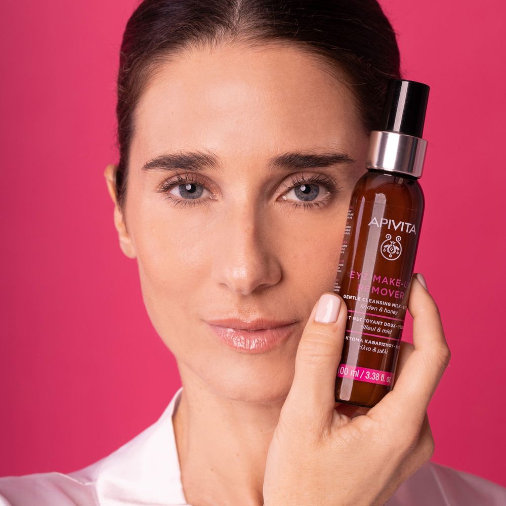 Donna tiene APIVITA Eye Make-up Remover. Sfondo rosa. Sorride. Pelle chiara, occhi blu.