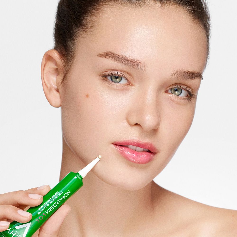Jeune femme tenant un tube vert. Inscription: Normaderm S.O.S. Anti-Pickel Sulfur Paste. L'embout touche la peau.