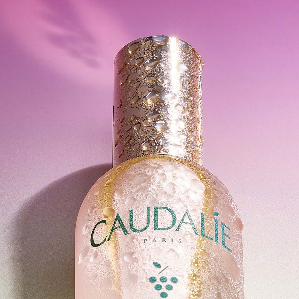 Flacone Caudalie Eau de Beauté, tappo argentato con gocce d'acqua. Testo Caudalie, uva. Sfumatura rosa-verde.