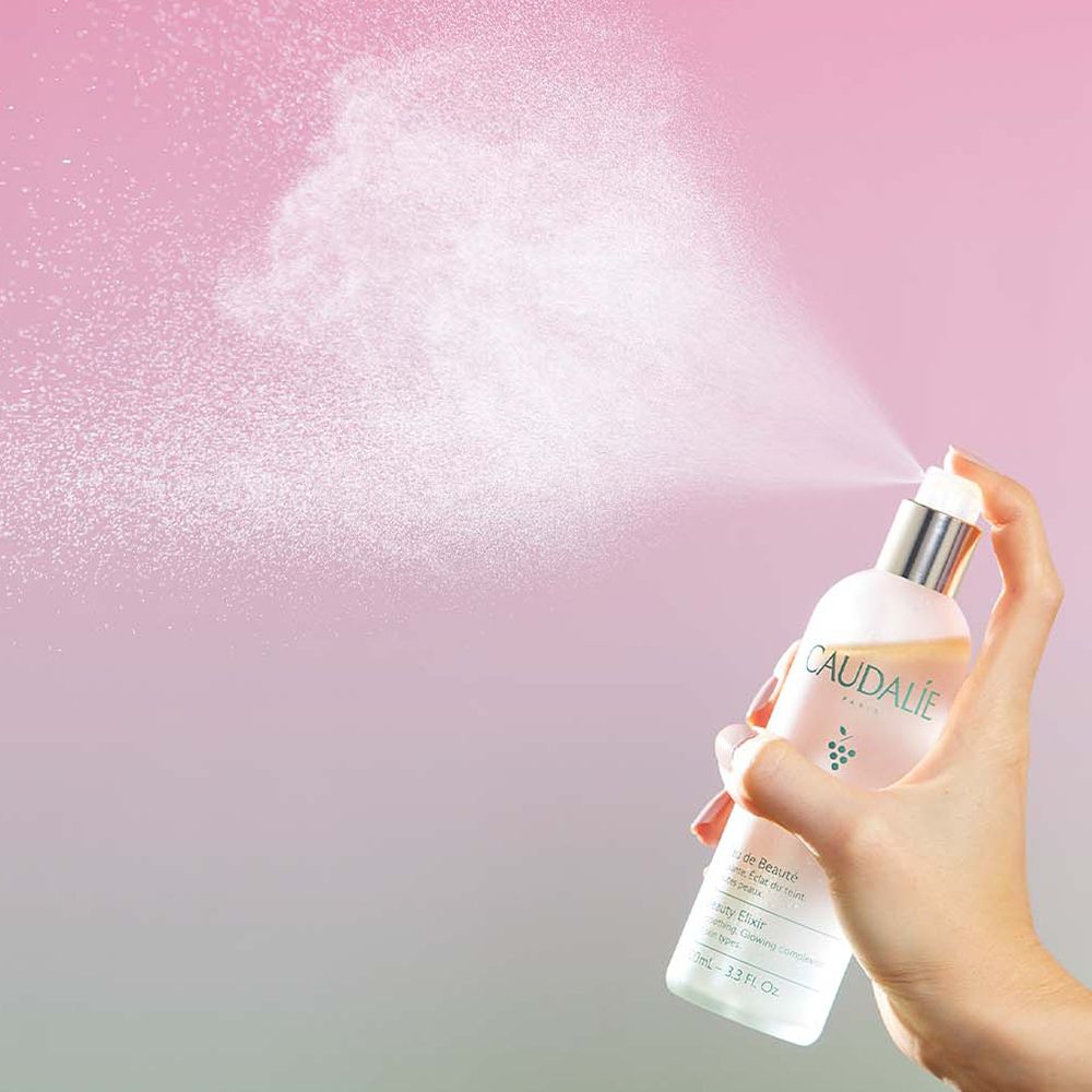 Mano che tiene flacone Caudalie Eau de Beauté. Nebulizzazione. Tappo argentato, sfumatura rosa-verde.