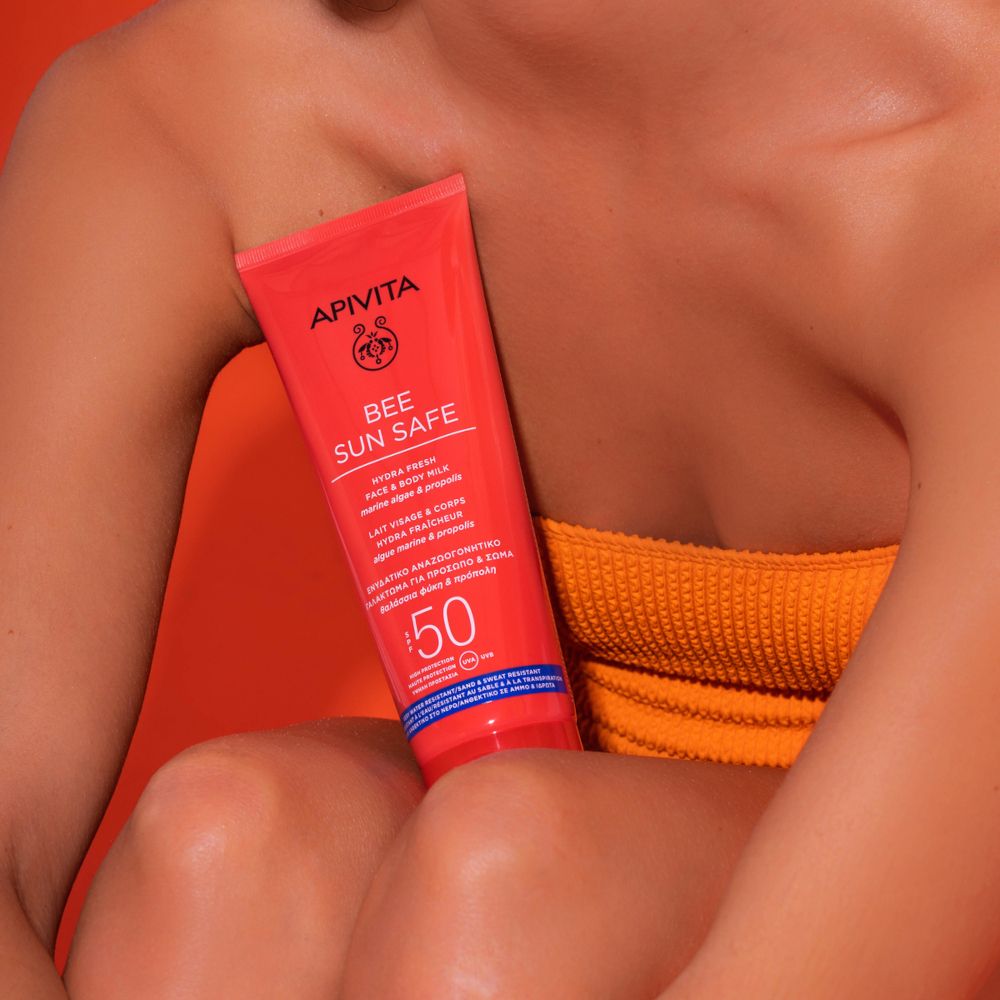 Donna con top arancione tiene un tubo rosso APIVITA BEE SUN SAFE. Testo: Hydra Fresh Face & Body Milk SPF50.