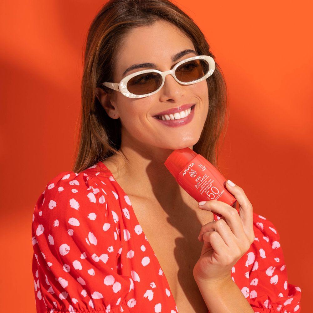 Donna con occhiali da sole tiene in mano un flacone rosso. Scritta: APIVITA, BEE SUN SAFE, SPF 50. Abito rosso.