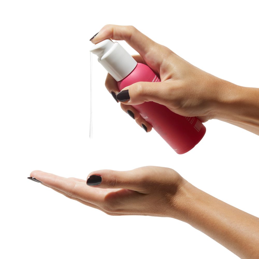 Mani che tengono flacone rosa con erogatore bianco. Liquido spruzzato nel palmo.
