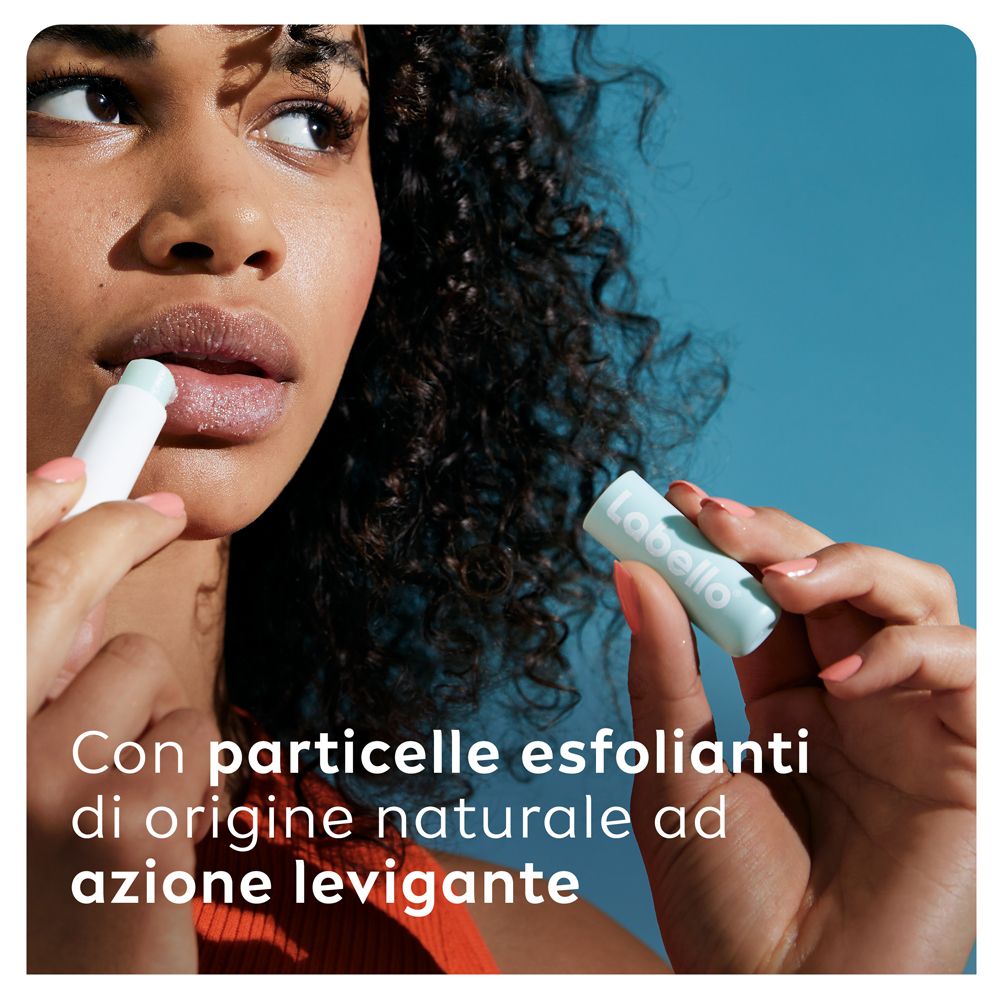 Donna usa Labello Caring Scrub. Prodotto in mano, applicazione sulle labbra. Testo: Con particelle esfolianti.