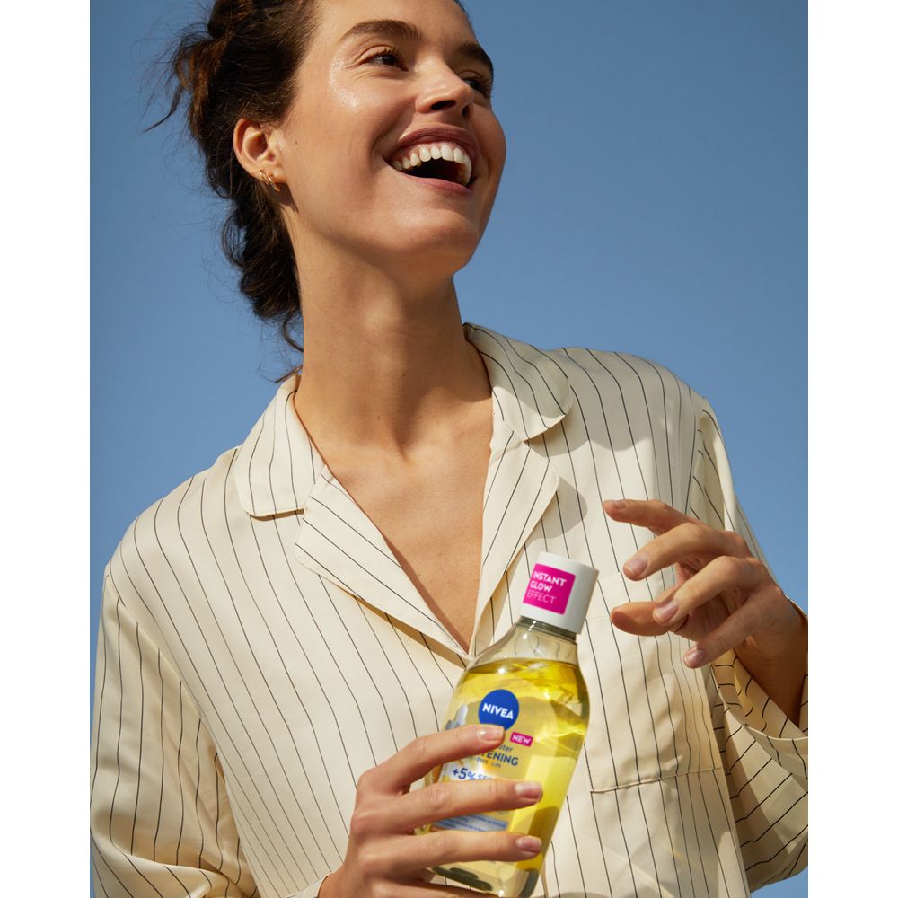 Donna sorridente tiene un flacone. Flacone con logo blu. Camicia a righe. Cielo blu.