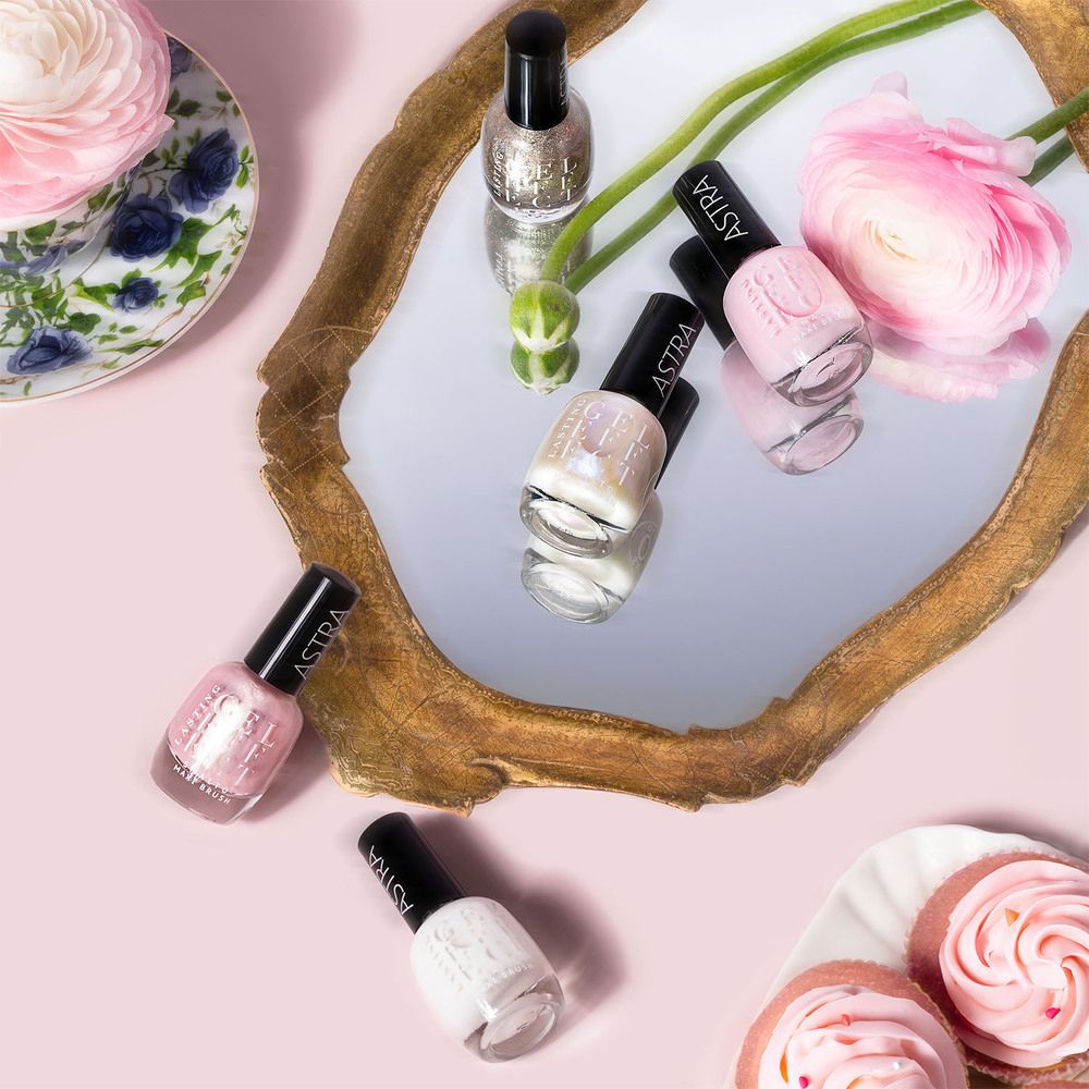 Diversi flaconi di smalto su uno specchio, circondati da fiori e pasticcini. I flaconi hanno tappi neri.