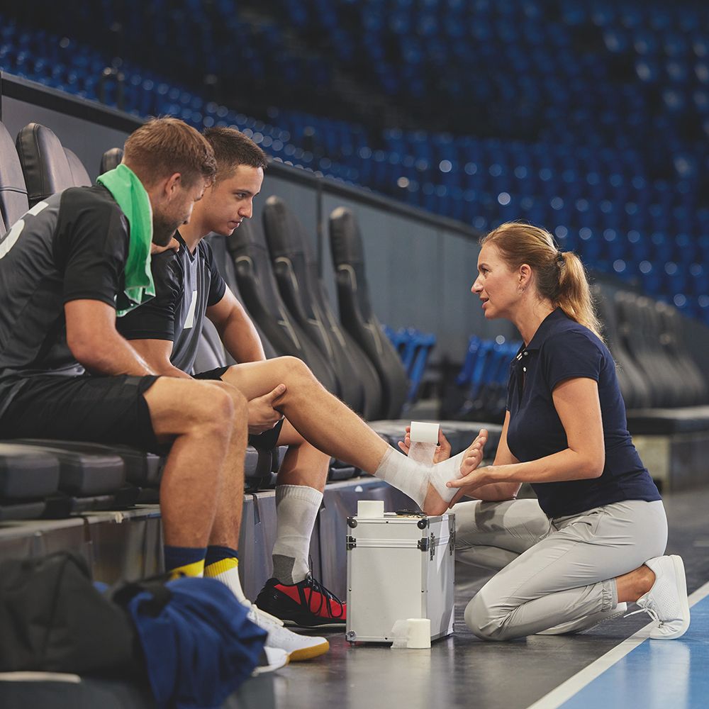 Sportler mit Bandage am Fuß. Frau wickelt Verband. Weitere Sportler sitzen daneben.