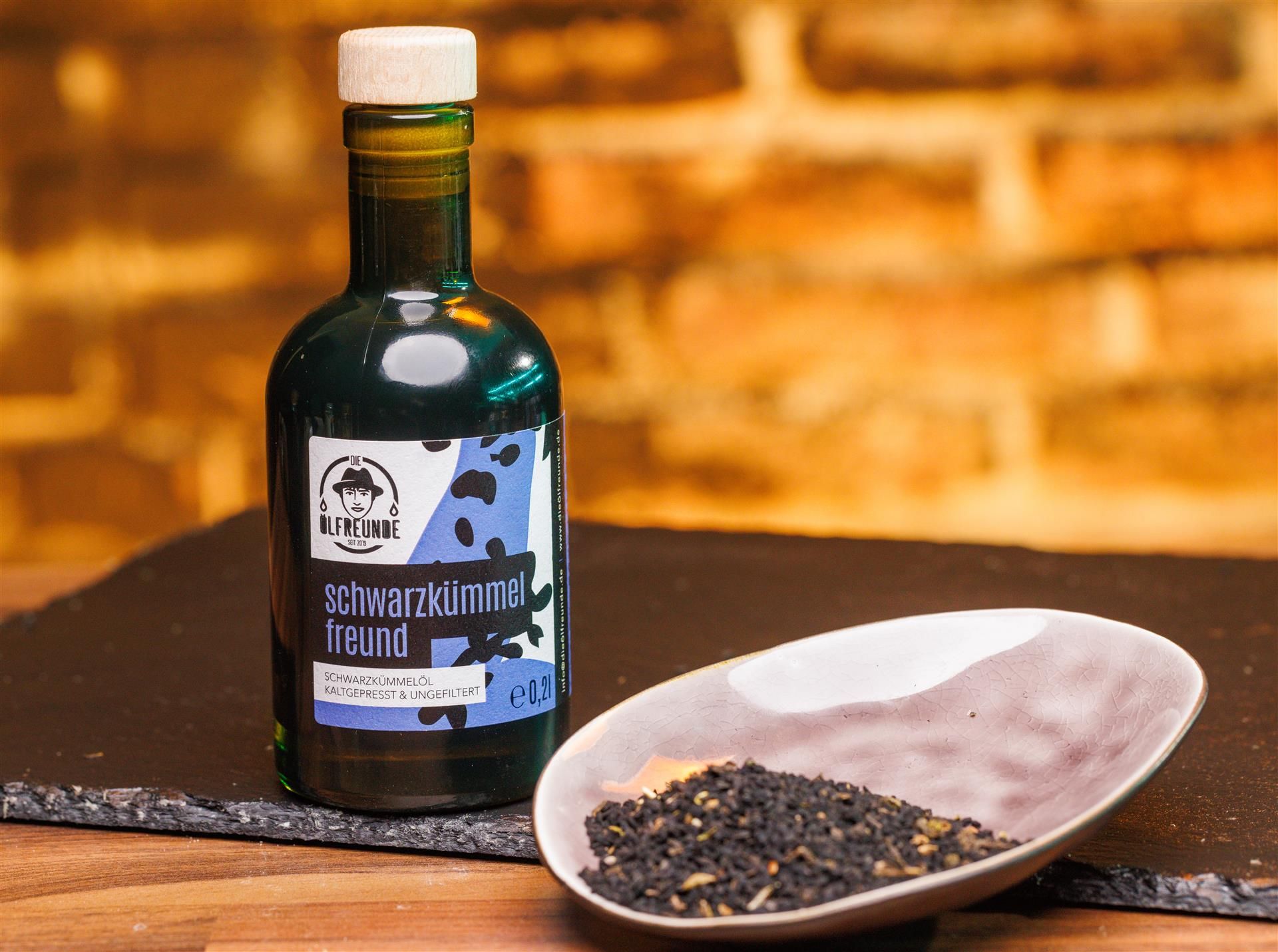 Braune Glasflasche mit Holzverschluss und Schale mit Samen. Etikett mit Produktname und Logo. Schwarzkümmelöl.