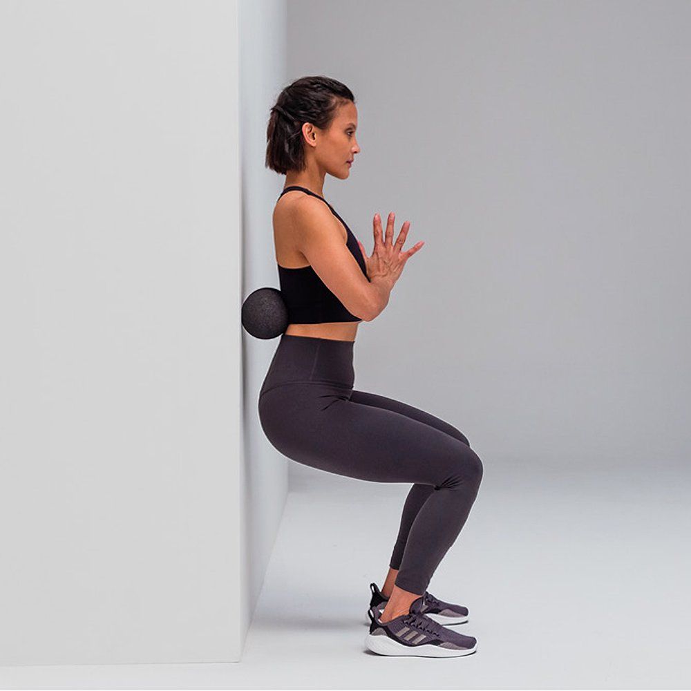 Person in Kniebeuge mit schwarzem Ball am Rücken. Wand im Hintergrund. Sportkleidung.