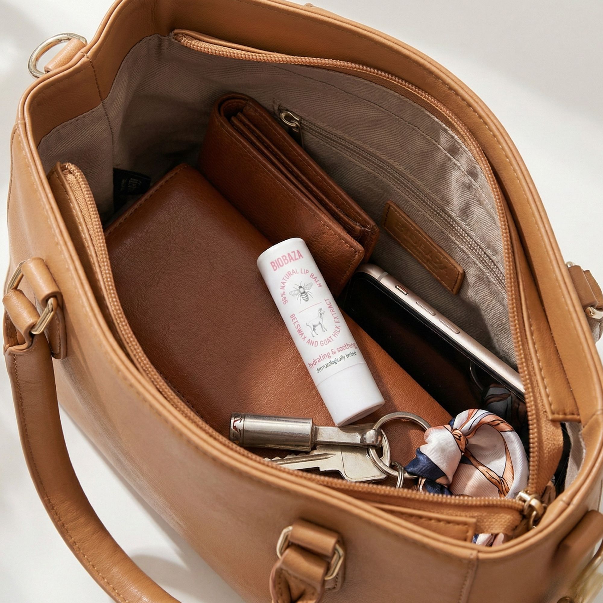 Offene Handtasche mit Lippenbalsam-Stift, Geldbörsen, Handy und Schlüsseln. Aufschrift: Biobaza. Produkt: Lippenbalsam-Stift.