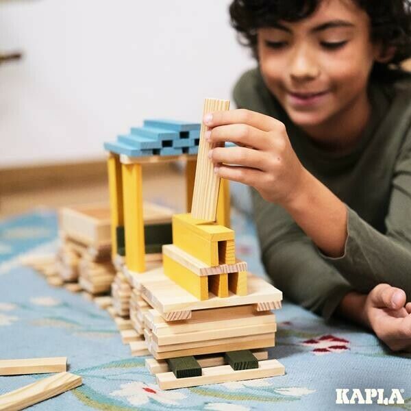 Kind baut Zug aus Holzplanken. Einige Planken sind blau und gelb gefärbt. Aufschrift: KAPLA.