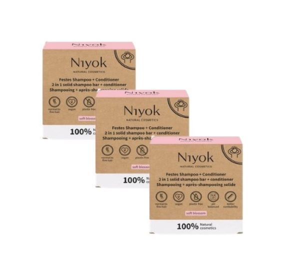 Drei Kartonverpackungen von Niyok 2-in-1 festes Shampoo + Conditioner. Die Verpackungen sind aus braunem Karton mit rosa Deckel. Auf der Vorderseite steht "Niyok Natural Cosmetics" und "Festes Shampoo + Conditioner". Es gibt auch Logos für "natürlich", "vegan" und "tierversuchsfrei".