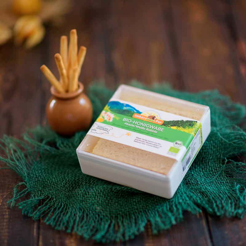 Bio-Honigwabe in Verpackung auf grünem Tuch. Daneben ein kleiner Krug mit Brotstangen. Bio-Siegel sichtbar.