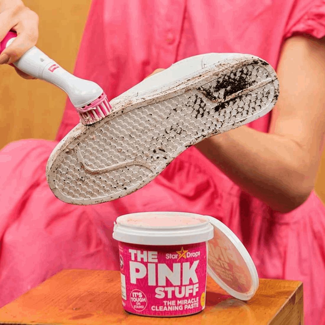 THE PINK STUFF Sonic Scrubber reinigt Schuhsohle. Daneben Tiegel THE PINK STUFF Reinigungs-Paste. Person trägt rosa Kleid.