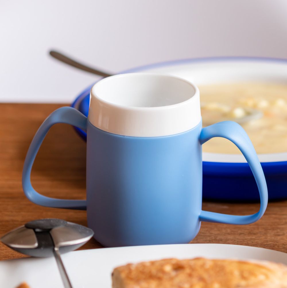 Blauer Becher mit zwei Griffen und weißem Rand. Auf einem Tisch, neben Essen.