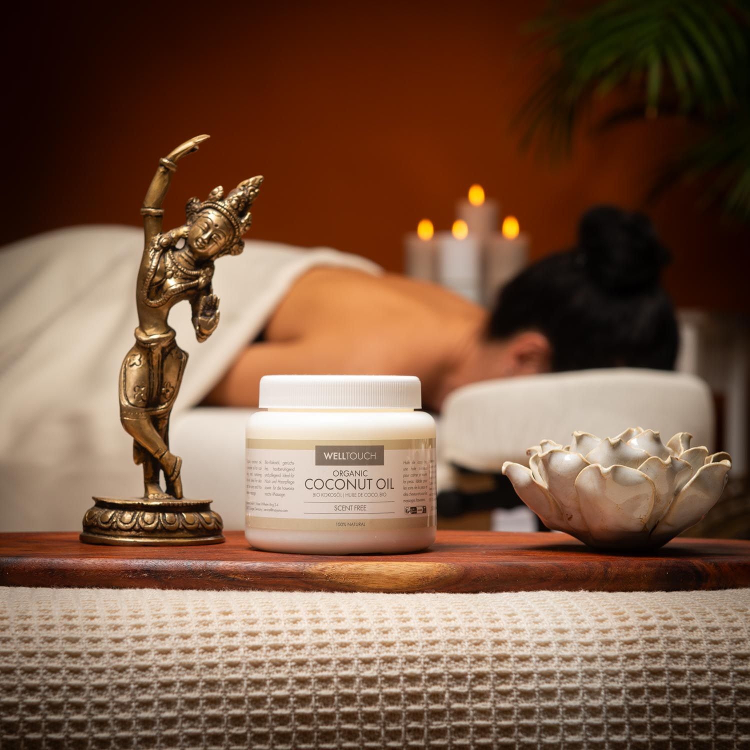Weißes Glas mit Aufschrift COCONUT OIL. Person erhält Massage. Bronze-Figur und Lotusblüte auf Holzbrett.