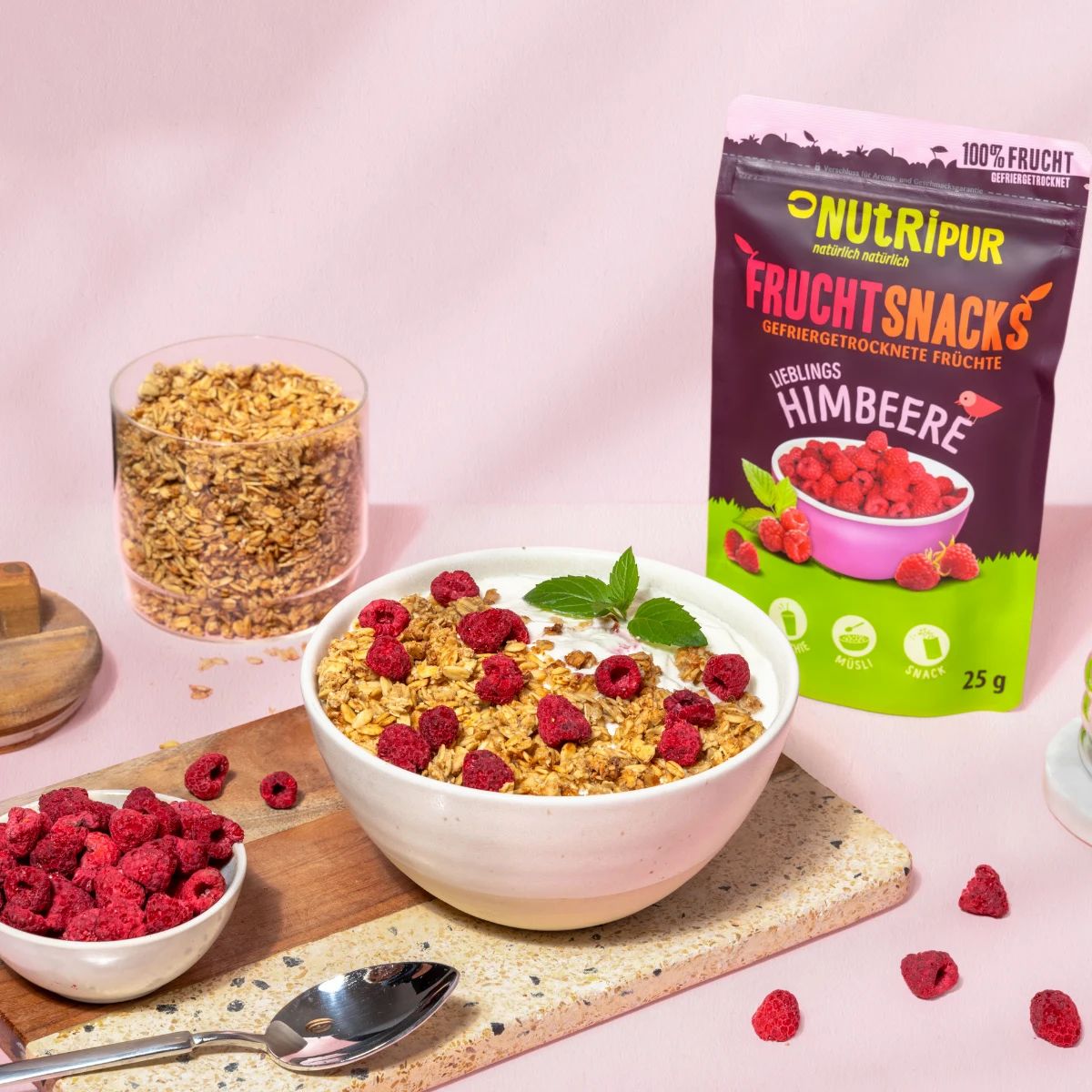 Schüssel mit Müsli, Himbeeren und Minze. Daneben Beutel NutriPur Fruchtsnacks Himbeeren. Glas mit Müsli.
