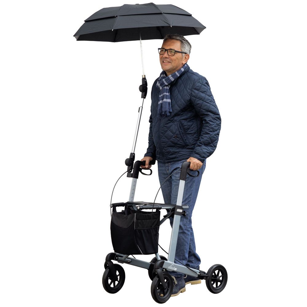 Ein Mann mit Brille und Schal steht neben einem Rollator mit aufgespanntem schwarzem Regenschirm.