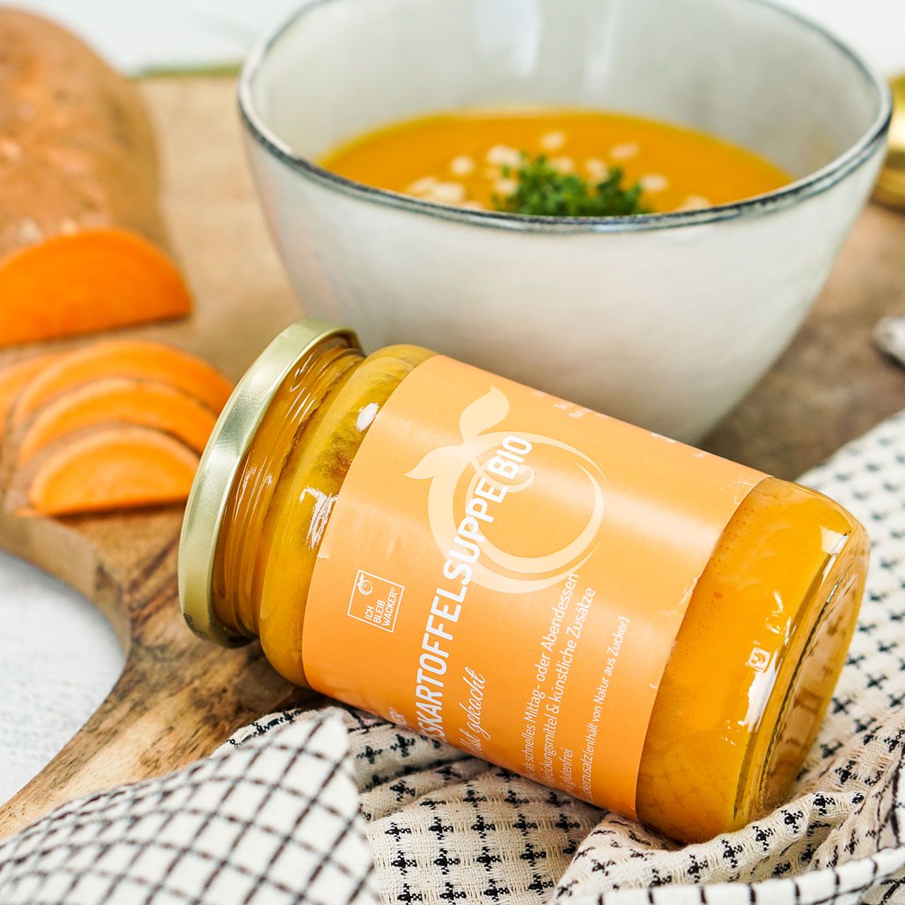 Glas Wacker Süßkartoffelsuppe Bio, liegend. Auf Holzbrett, neben Schüssel mit Suppe, Karottenscheiben.