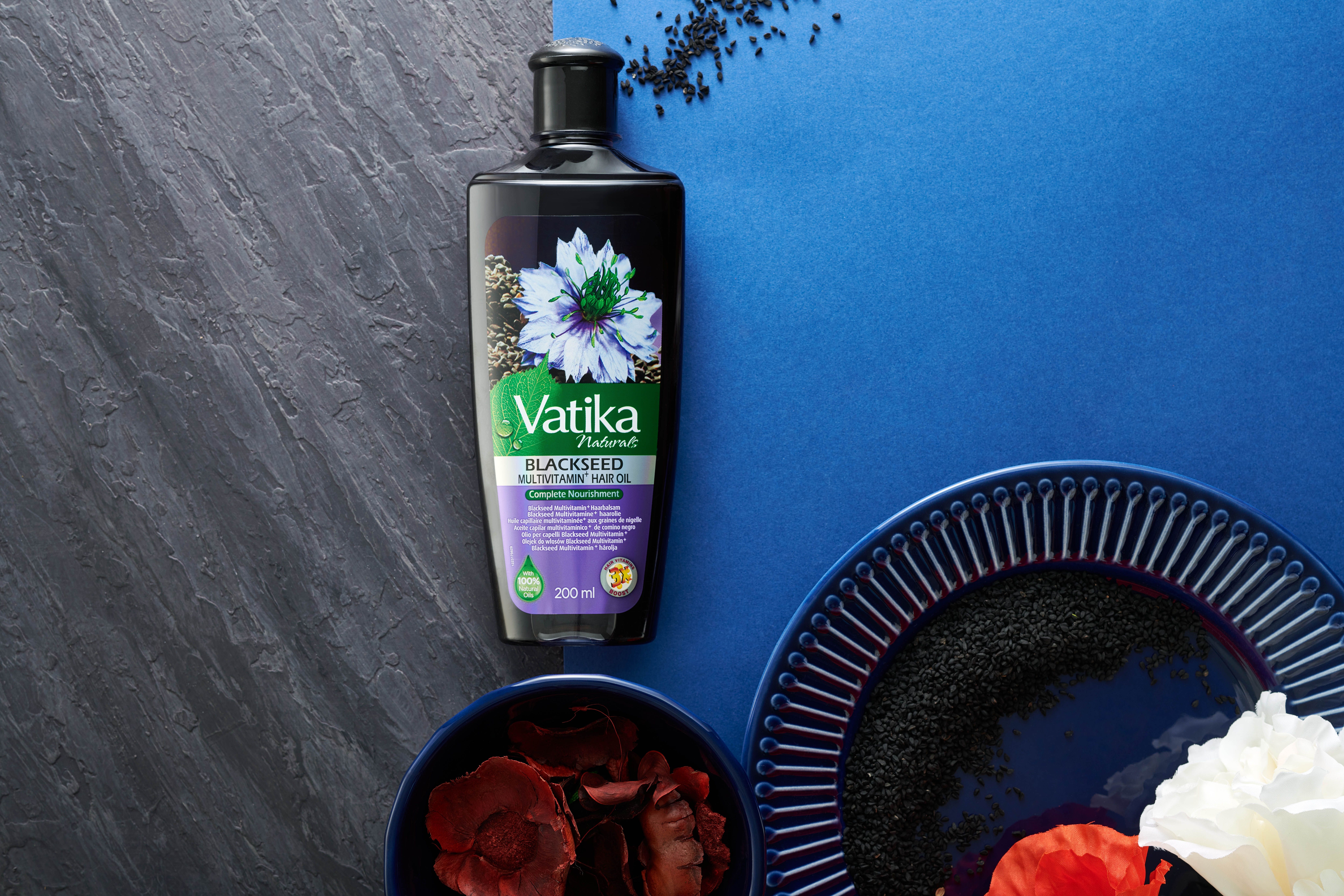 Vatika Naturals Schwarzkümmel Haaröl auf blauem Untergrund. Neben Schalen mit Zutaten und einem Teller.