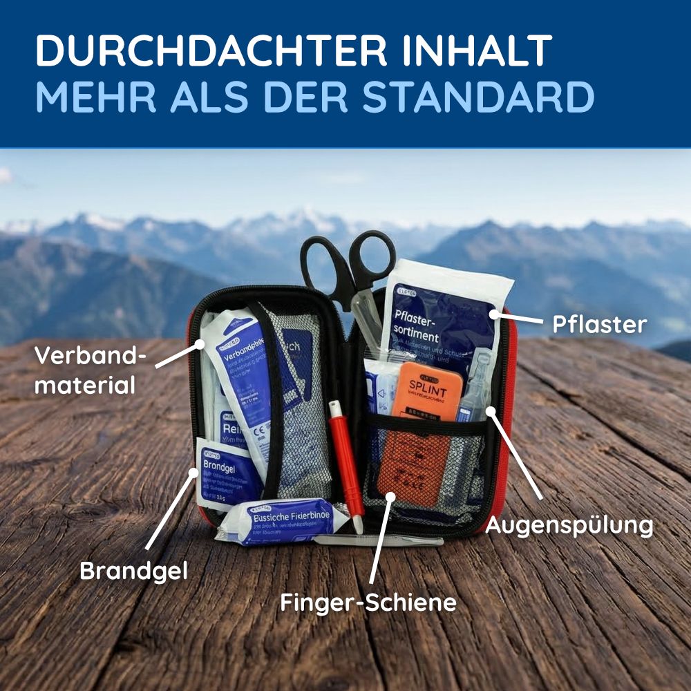 Geöffnetes Erste-Hilfe-Set mit Verbandsmaterial, Pflastern, Brandgel, Augenspülung und Finger-Schiene. Auf einem Tisch mit Bergpanorama im Hintergrund.