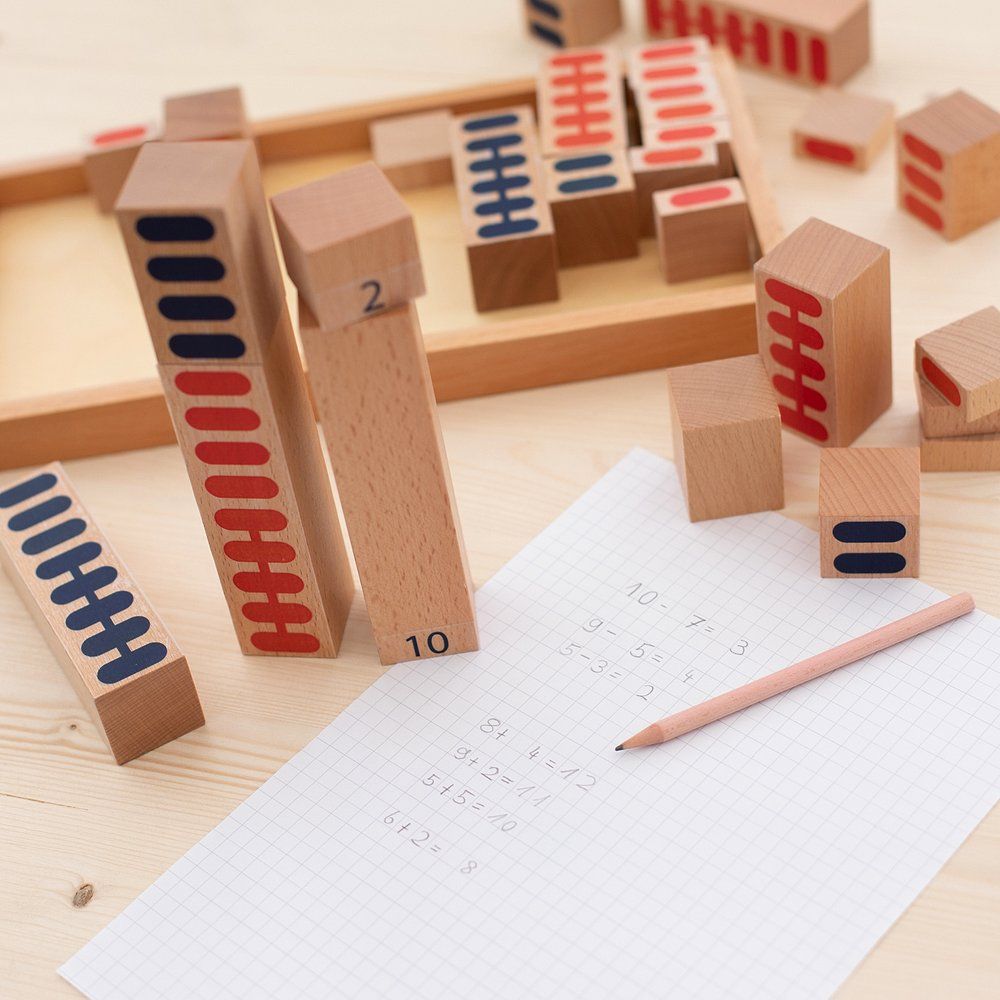 Holzbausteine mit Zahlen und farbigen Markierungen. Einige Bausteine liegen auf einem Tisch. Ein Blatt Papier mit Bleistift und Rechenaufgaben.
