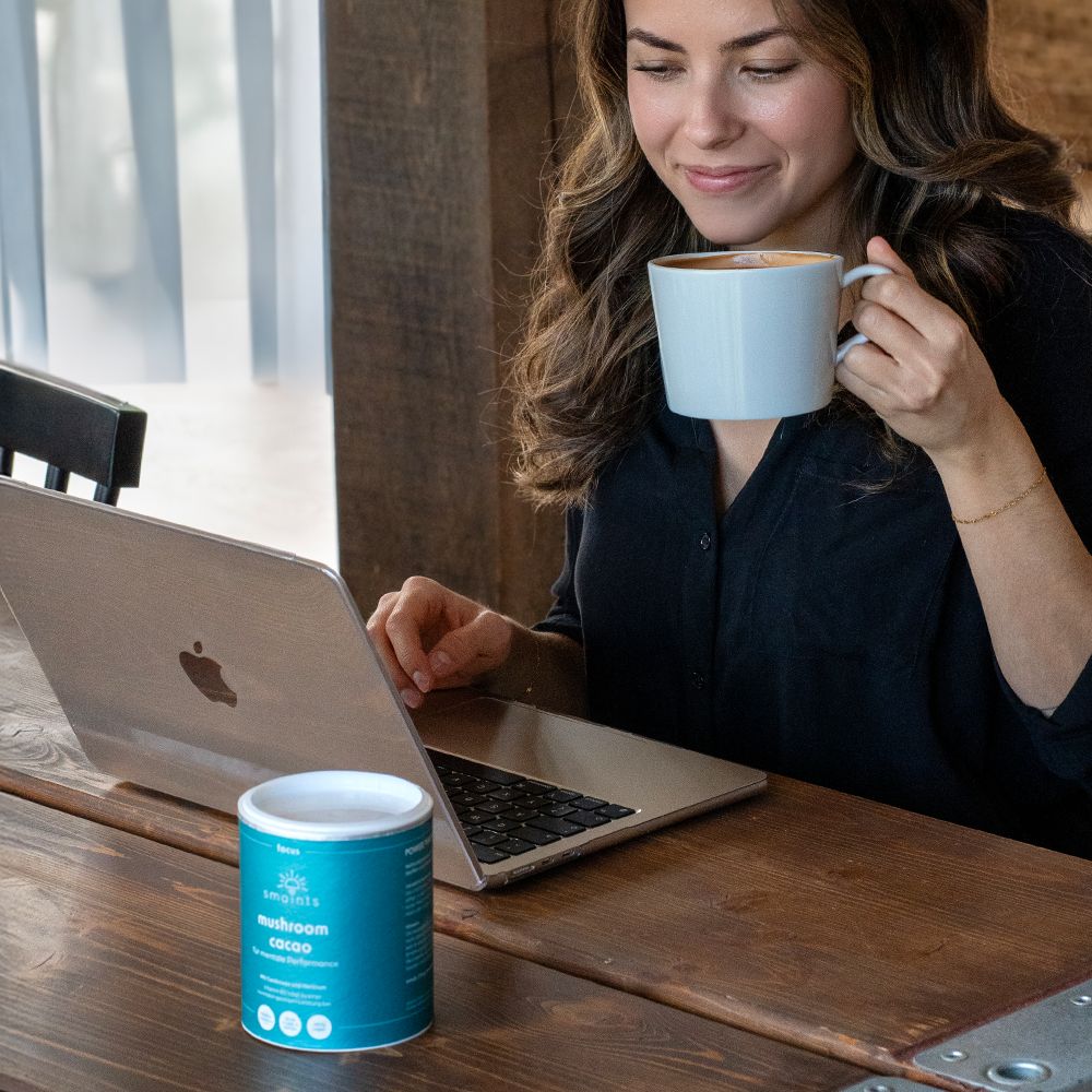 Frau trinkt aus einem weißen Becher. Auf einem Tisch steht eine Dose. Laptop auf dem Tisch.