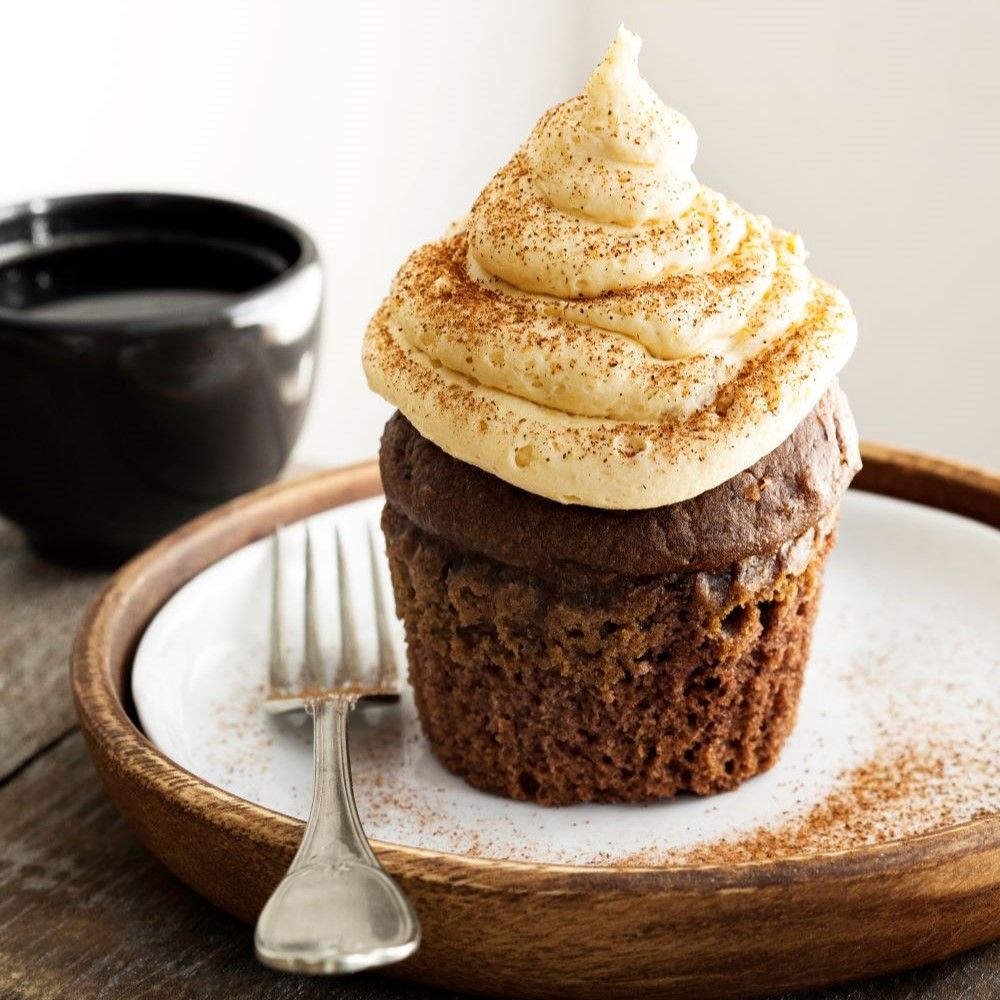 Cupcake mit cremiger Glasur und Zimt bestreut. Auf einem Teller mit Gabel. Schwarze Tasse im Hintergrund.