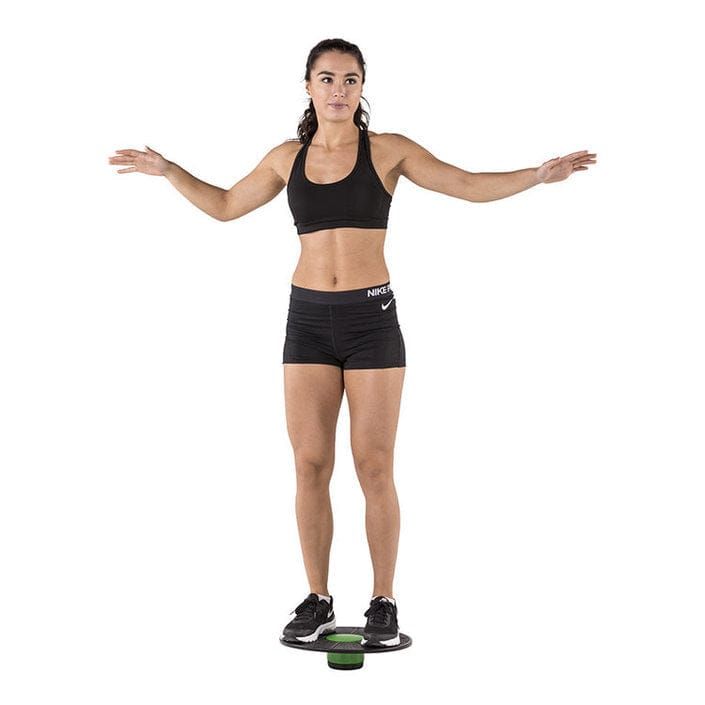 Frau steht auf Balanceboard. Schwarze Sportkleidung. Arme seitlich ausgestreckt. Balanceboard mit grünem Element.